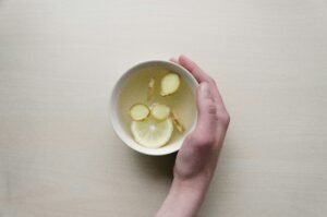 A mug of water with few lemon and ginger wedges, and a hand holding the mug.