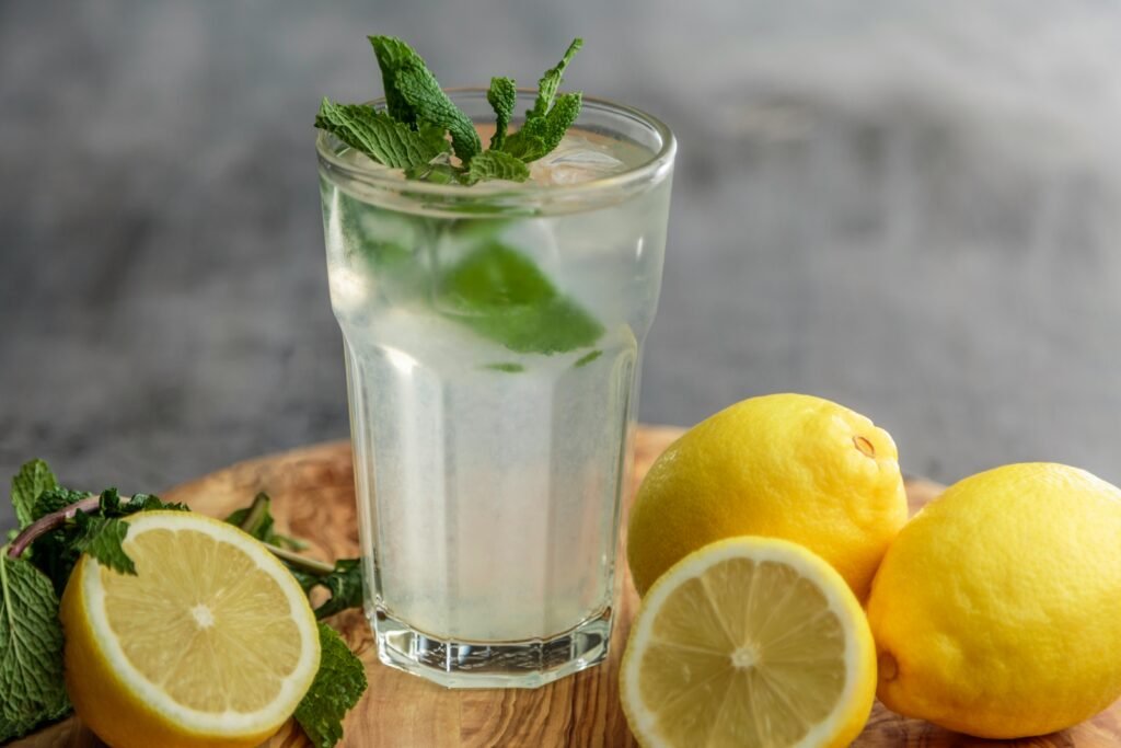 A glass of lemon water topped with mint leaves and next to it there are few lemons spread on the table.